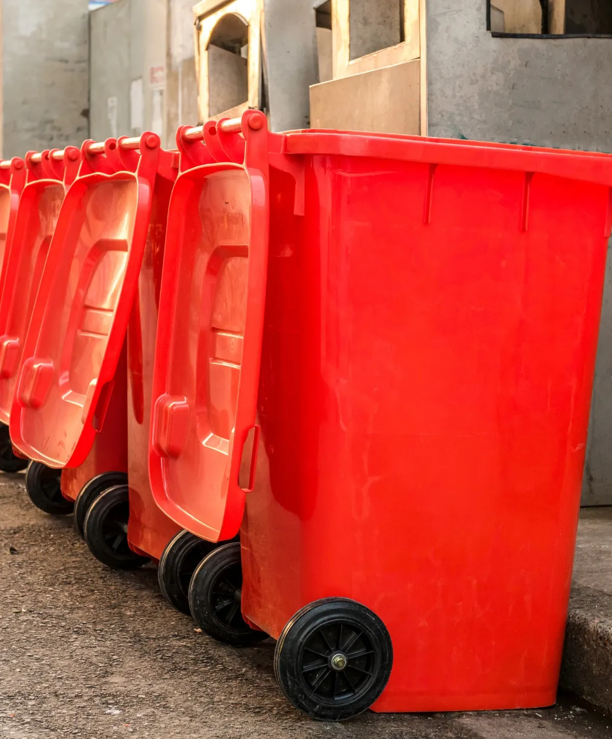 A row of several bright red wheeled trash bins with their lids open, lined up on a street next to a building.