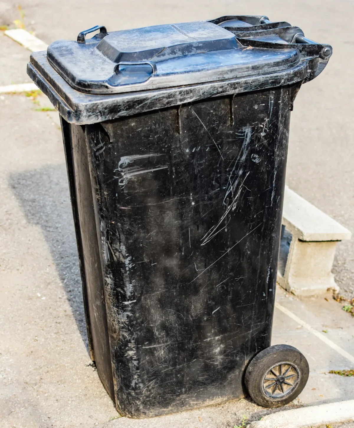 A weathered, scratched black wheeled garbage bin with its lid closed stands on a paved surface outdoors.