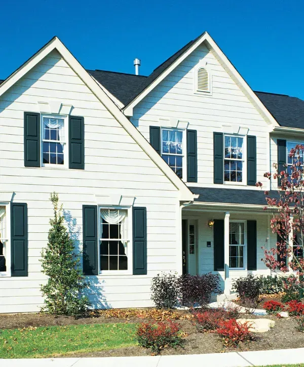 White suburban home with black shutters and a manicured front yard with bushes and a walkway. 