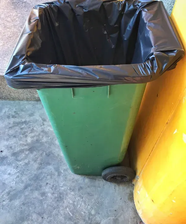 Green trash bin with a black garbage bag liner placed next to a yellow wall on a concrete floor.
