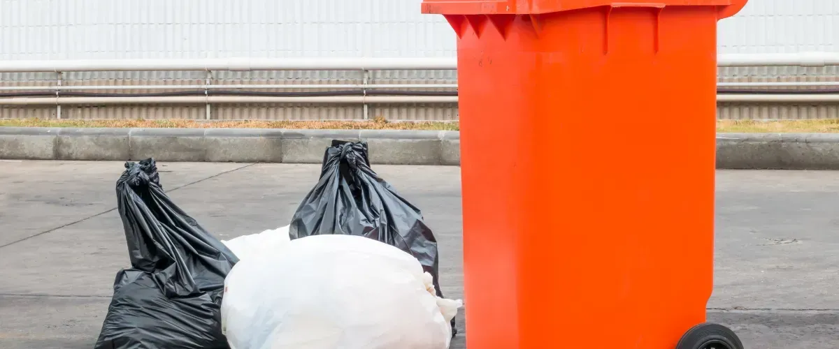 Black and white trash bags placed on the ground beside a large orange outdoor garbage bin.