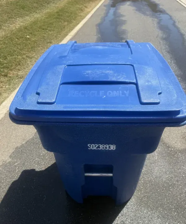 A top-down view of a blue recycling bin with "RECYCLE ONLY" on its lid, set on a driveway next to grass.