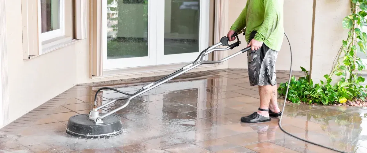 A person in a green shirt power washing a wet tiled patio with a surface cleaner tool, next to a beige building.