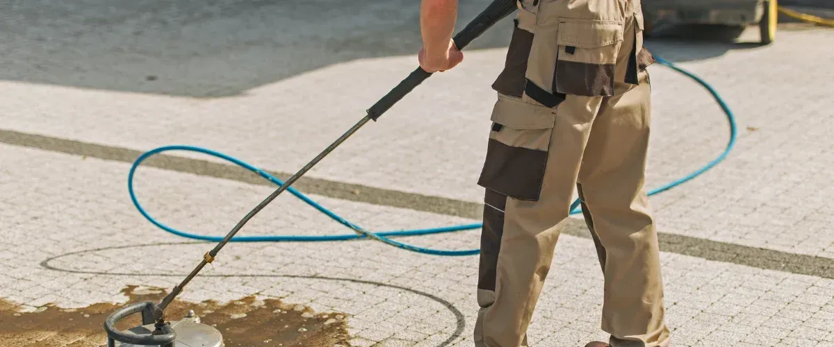 A person in khaki work pants uses a power washer to clean a dirty, light-colored brick pavement.