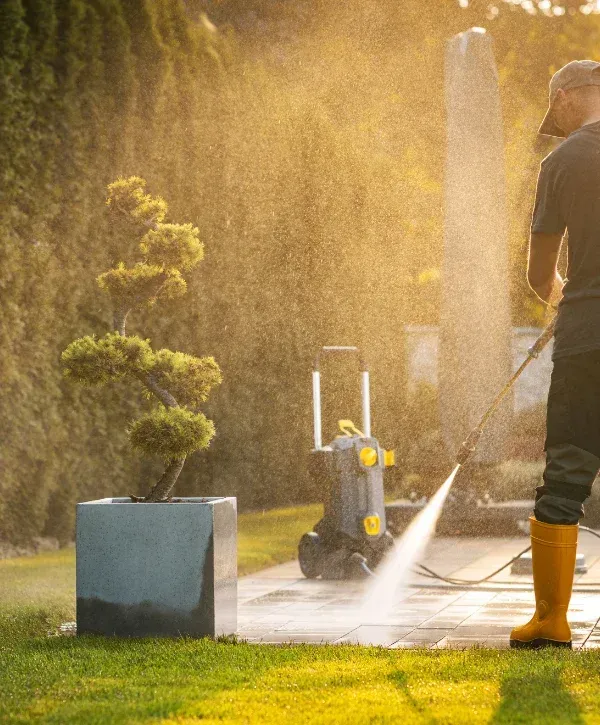 A man in yellow boots pressure washing a patio next to a bonsai tree in a concrete planter, with golden sunlight.