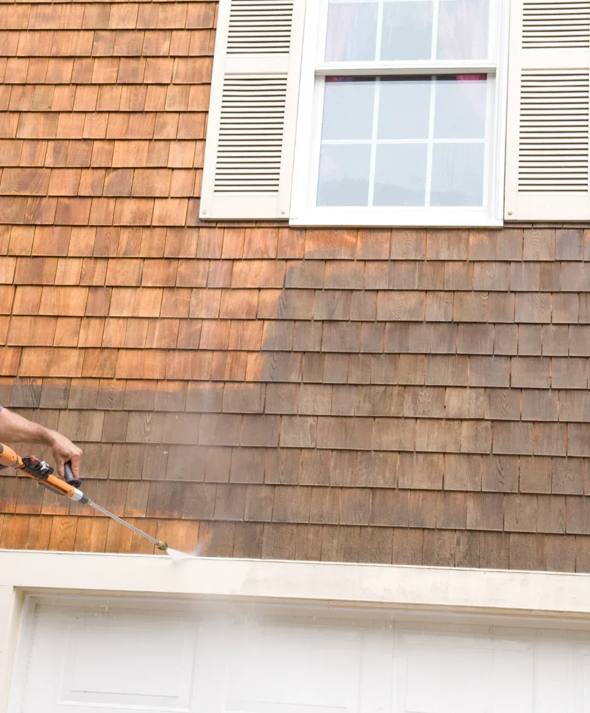 A person pressure washing cedar shake siding, showing a clean lighter section and a dirty darker section.