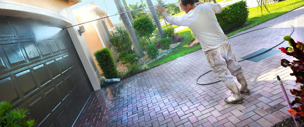 A man pressure washing a dirty concrete driveway, creating a clean wet strip next to dry, grimy pavement.