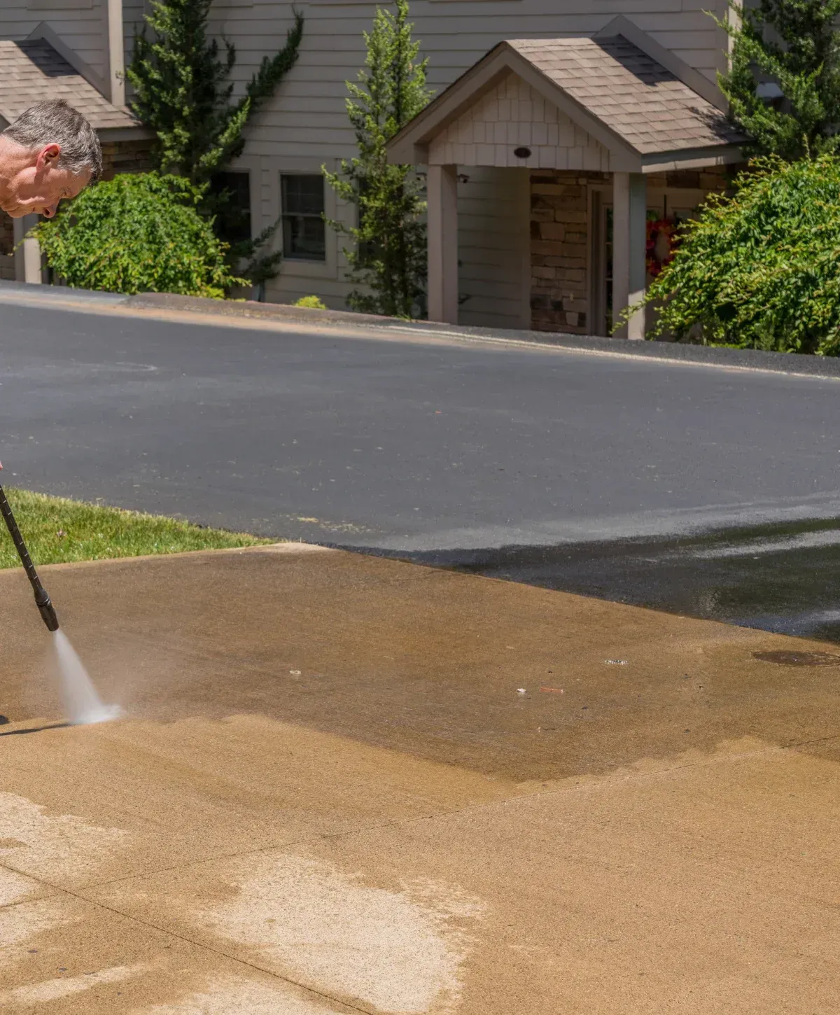 A man in dirty white clothes power washing a dark garage door and brick paver driveway on a sunny day.