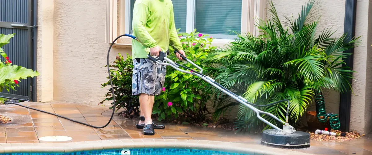 A person in a lime green shirt and camo shorts uses a surface cleaner to pressure wash a pool patio.
