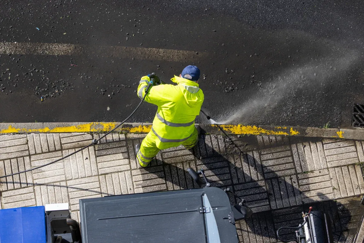 Overhead view of a person in a bright yellow suit pressure washing a yellow-painted curb separating a street and sidewalk.