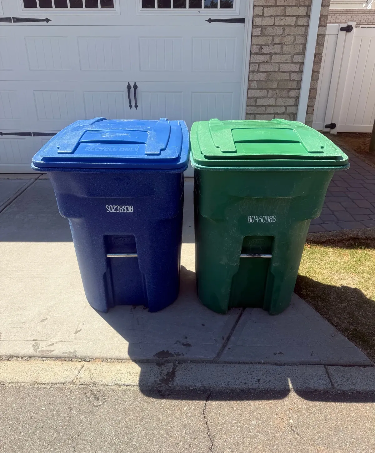 Two large residential bins, one blue for 'RECYCLE ONLY' and one green, stand side-by-side on a sunny driveway.