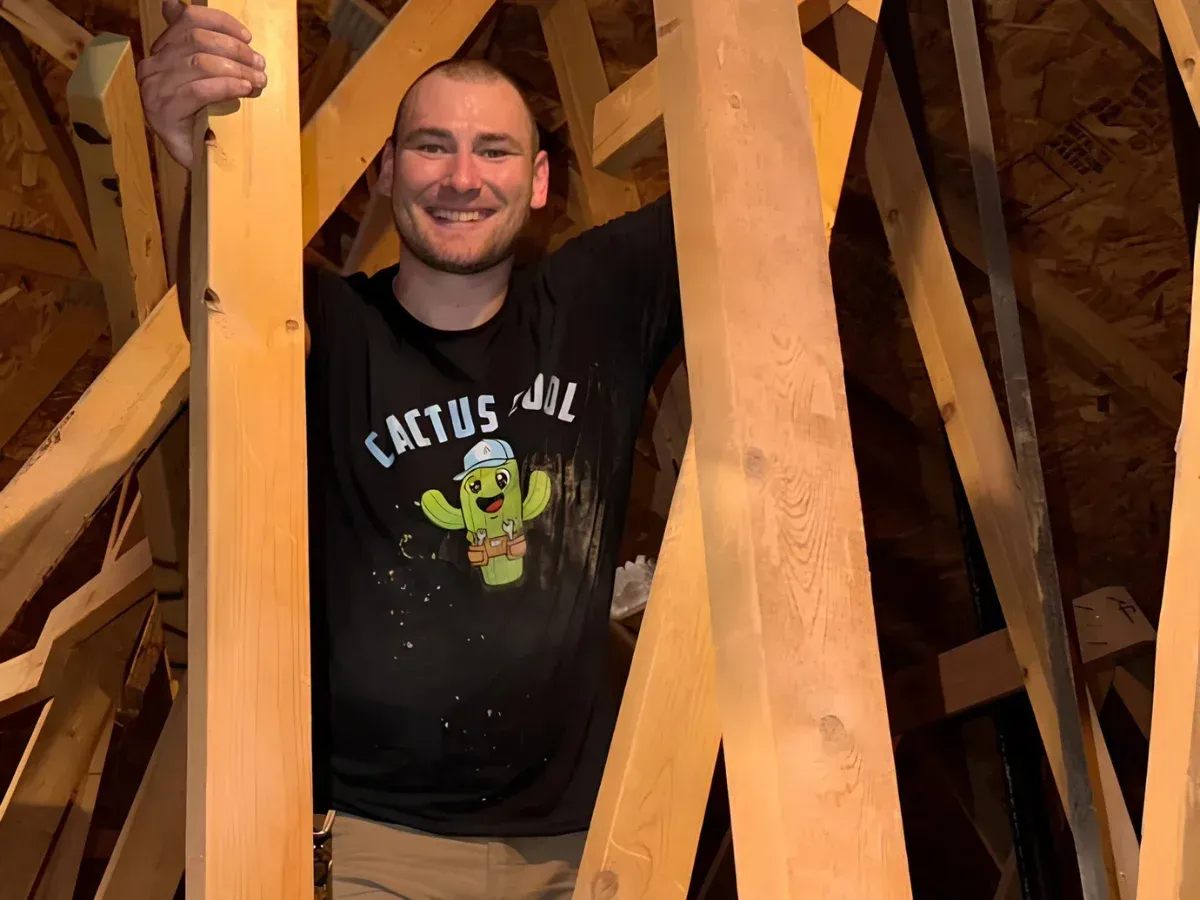 A man with a shaved head and a black 'Cactus Cool' t-shirt smiles in an attic surrounded by wooden beams.