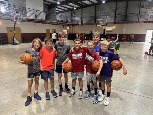 Two camp counselors, one male and one female, smiling and giving high-fives to a group of children in matching camp t-shirts. The setting is a shaded picnic area with colorful banners, evoking a sense of safety, mentorship, and community.