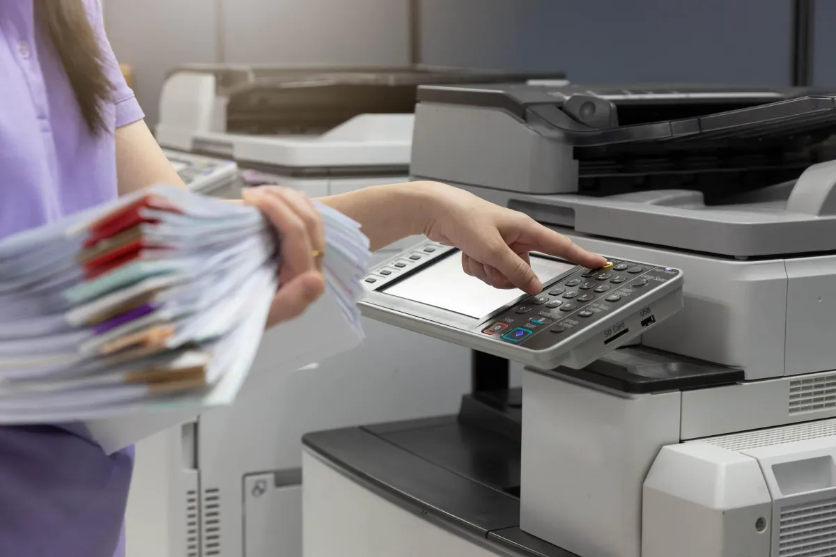An office worker holding a stack of files while operating the control panel of a high-volume copier