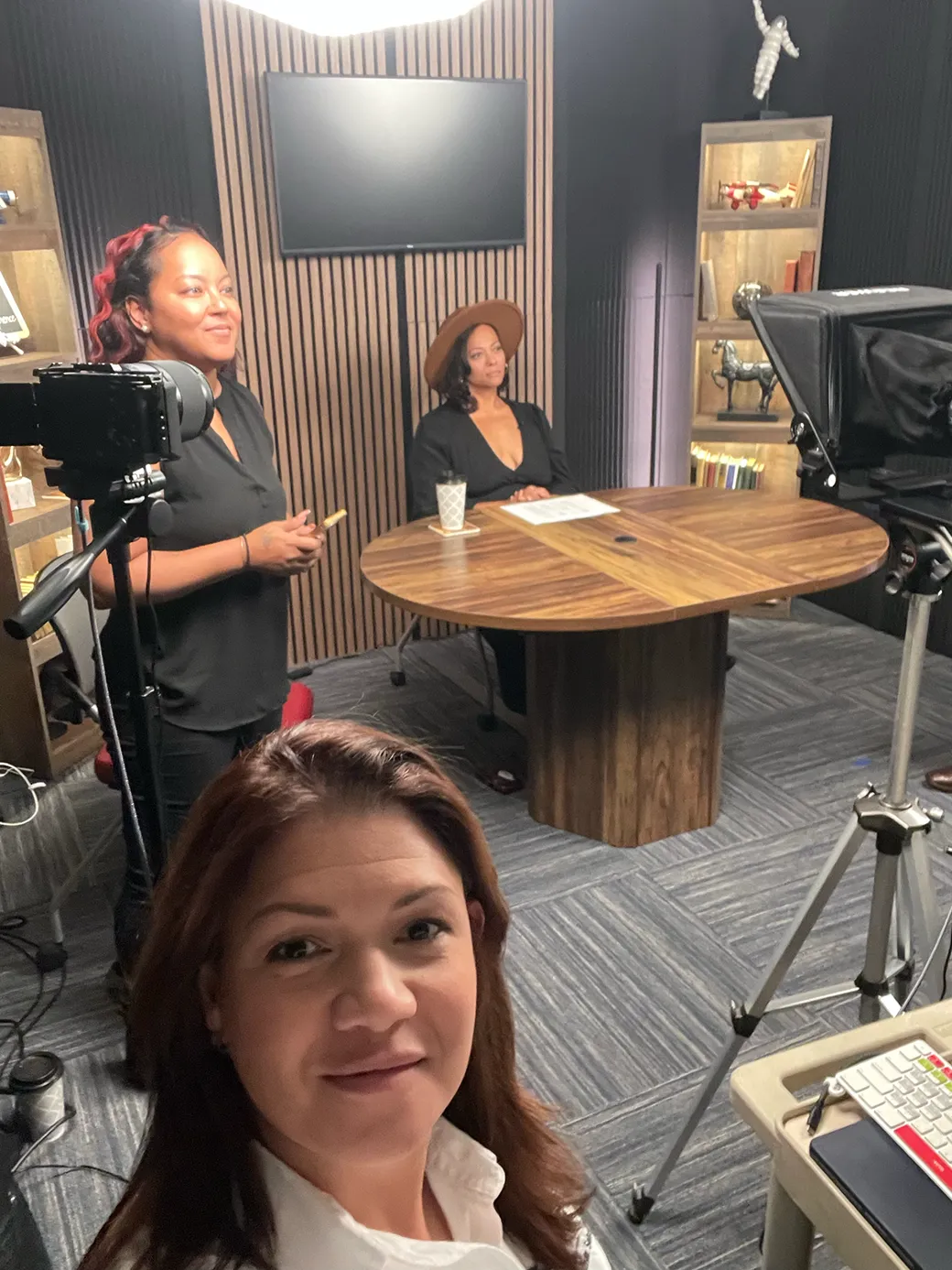 Professional podcast studio with acoustic panels, dual monitors, and a sleek mixing console. A Hispanic woman in headphones is recording, while a South Asian man adjusts audio levels. The room is high-tech, organized, and filled with creative tools.