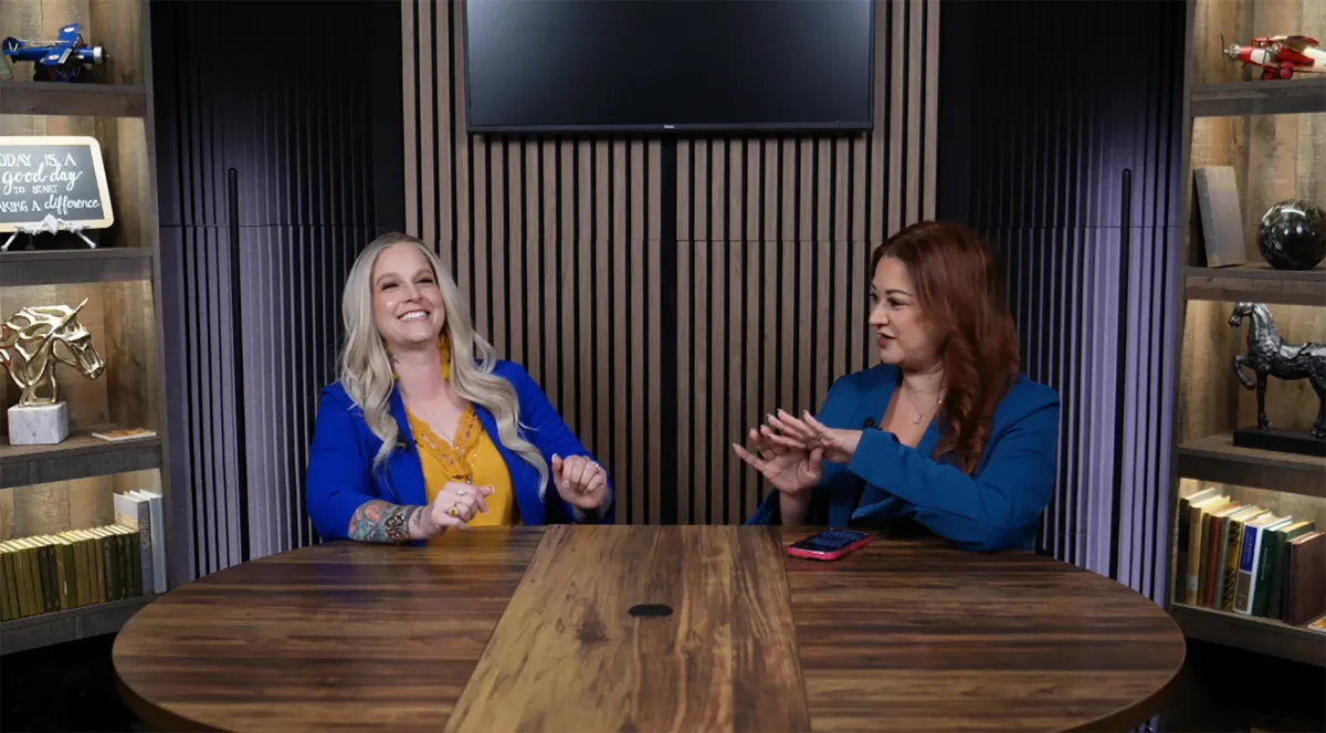 Professional podcast studio with acoustic panels, dual monitors, and a sleek mixing console. A Hispanic woman in headphones is recording, while a South Asian man adjusts audio levels. The room is high-tech, organized, and filled with creative tools.