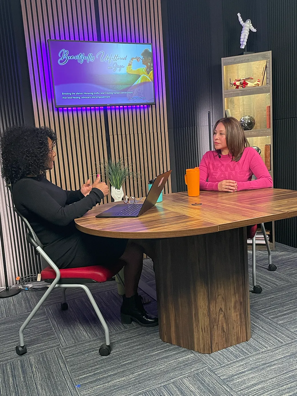 Professional podcast studio with acoustic panels, dual monitors, and a sleek mixing console. A Hispanic woman in headphones is recording, while a South Asian man adjusts audio levels. The room is high-tech, organized, and filled with creative tools.