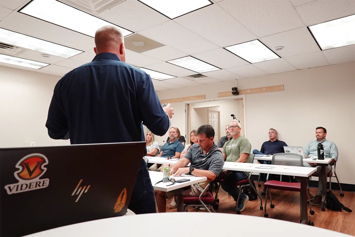 Modern, sunlit studio with glass walls, a large wooden table, professional microphones, and a city view. A diverse group of adults, including a Black woman and Asian man, are setting up for a podcast. The space is vibrant, organized, and exudes creative energy.