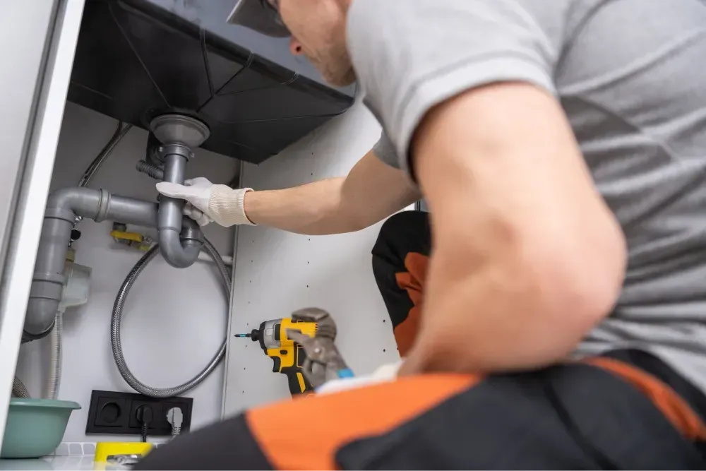 A plumber working under a kitchen sink using tools to clean and maintain drainage pipes.