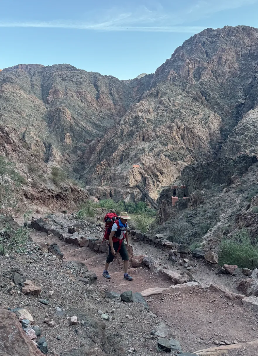 Karey Kosorok hiking a rocky trail in the Grand Canyon with a backpack, near the Colorado River suspension bridge.