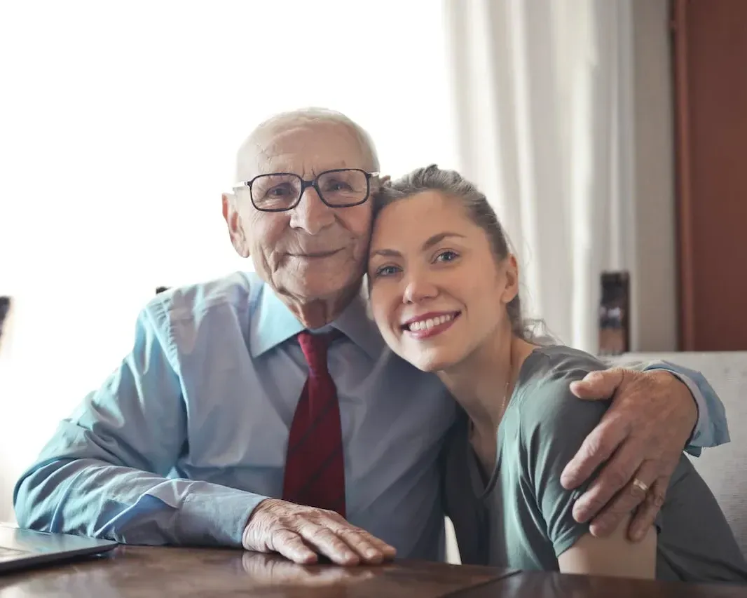 A photo of an elderly gentleman with arm over the shoulder of a younger lady both are smiling and looking at the camera