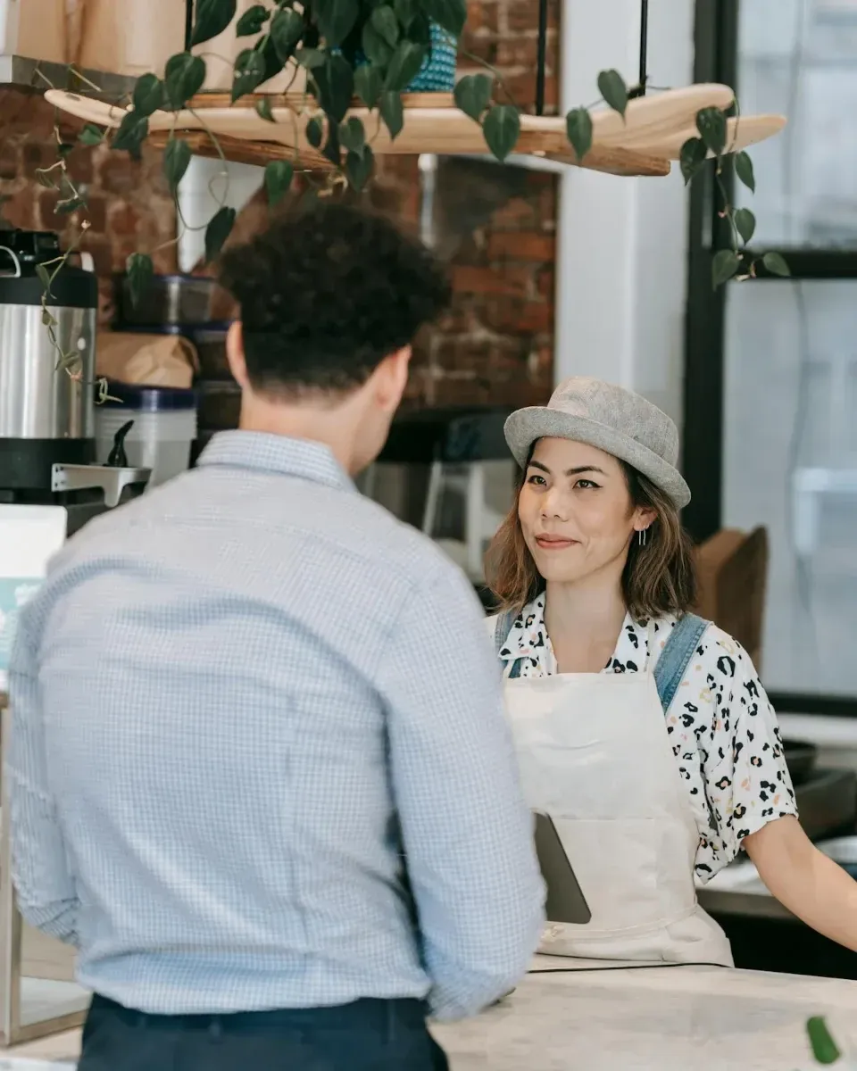 A photo of a young woman behind the cash register at coffee shop smiling at a customer approaching the counter