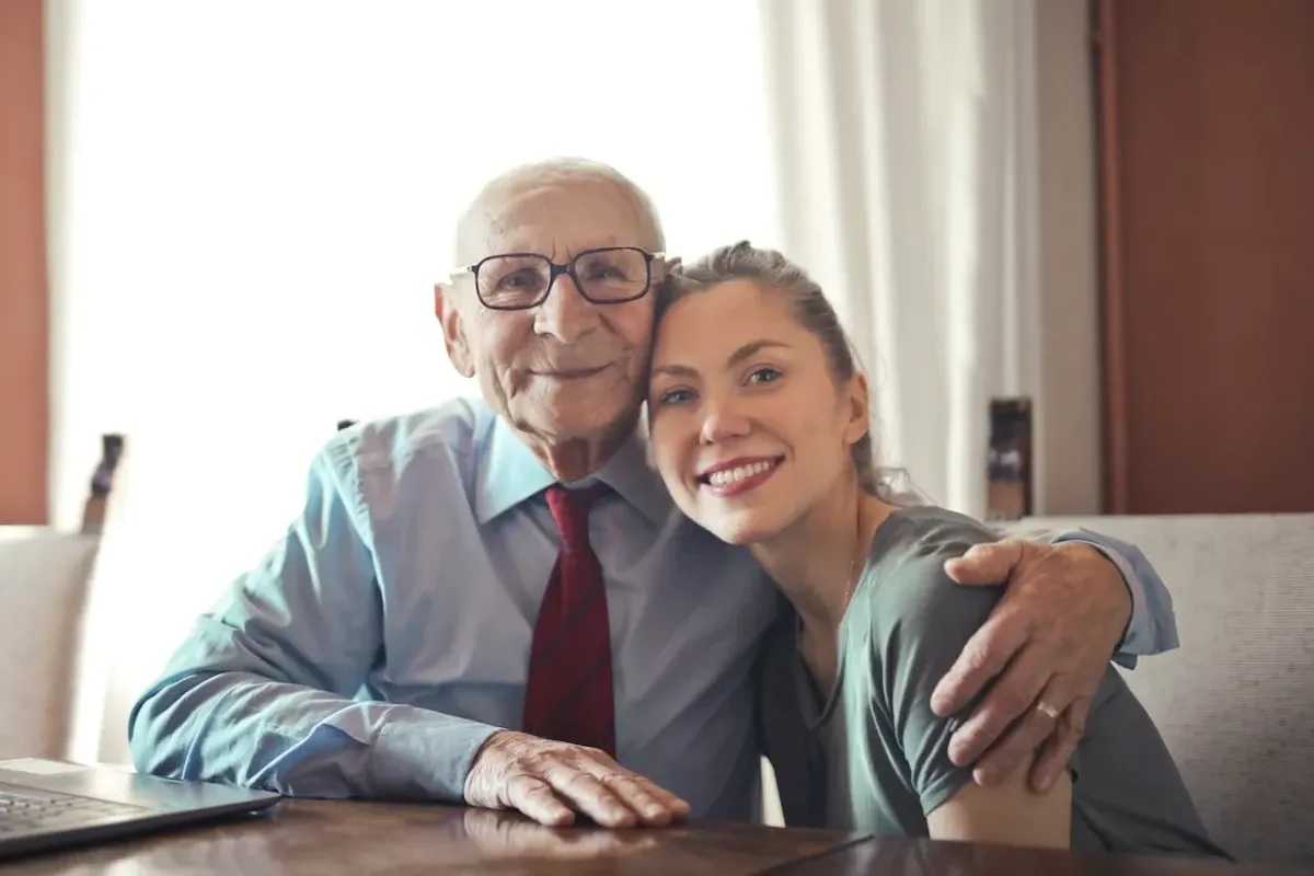 A photo of an elderly gentleman with arm over the shoulder of a younger lady both are smiling and looking at the camera