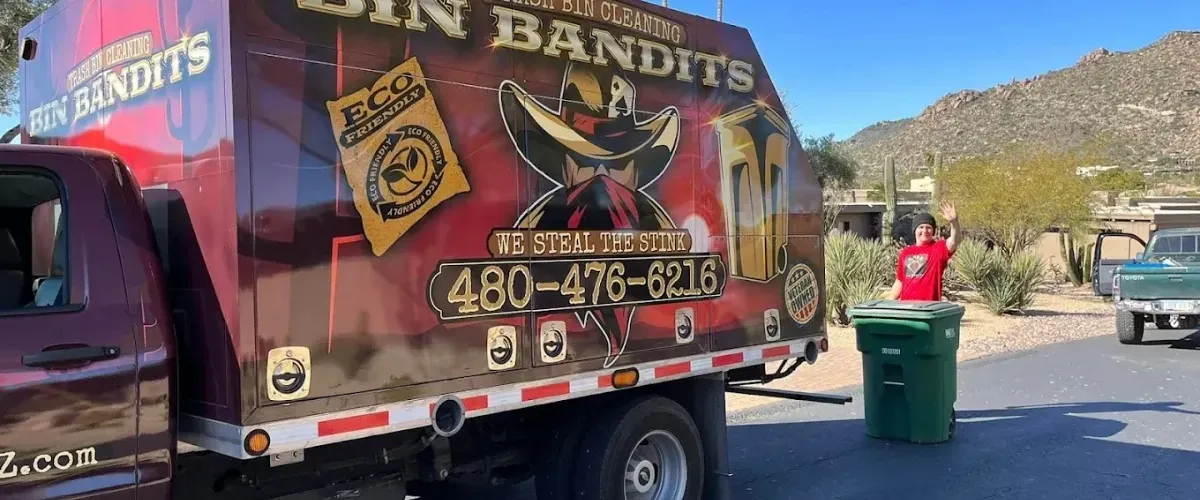 Bin Bandits trash bin cleaning truck operating in an Arizona neighborhood with a worker waving beside a green garbage bin.
