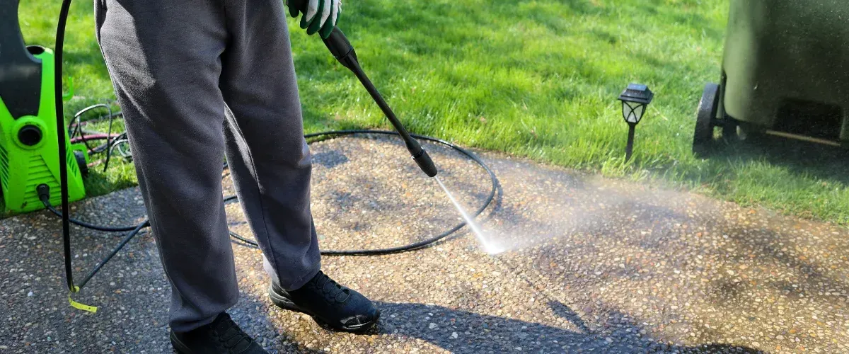 A person in gray sweatpants and gloves power washing a concrete pebble aggregate patio or path.