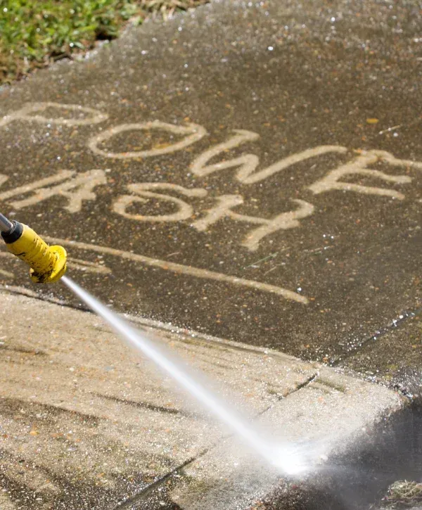 A person in a red uniform and green boots power washing a grimy stone sidewalk, with steam rising from the water.