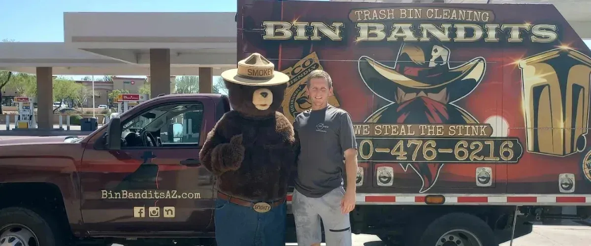 A man and Smokey Bear mascot stand before a Bin Bandits trash bin cleaning truck.