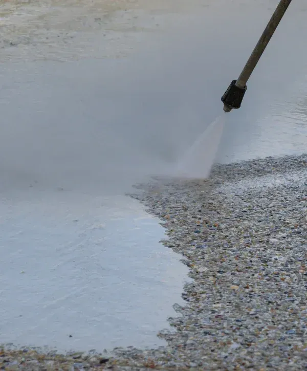A power washer stream cleaning a dirty, pebbled concrete surface, revealing a cleaner path.