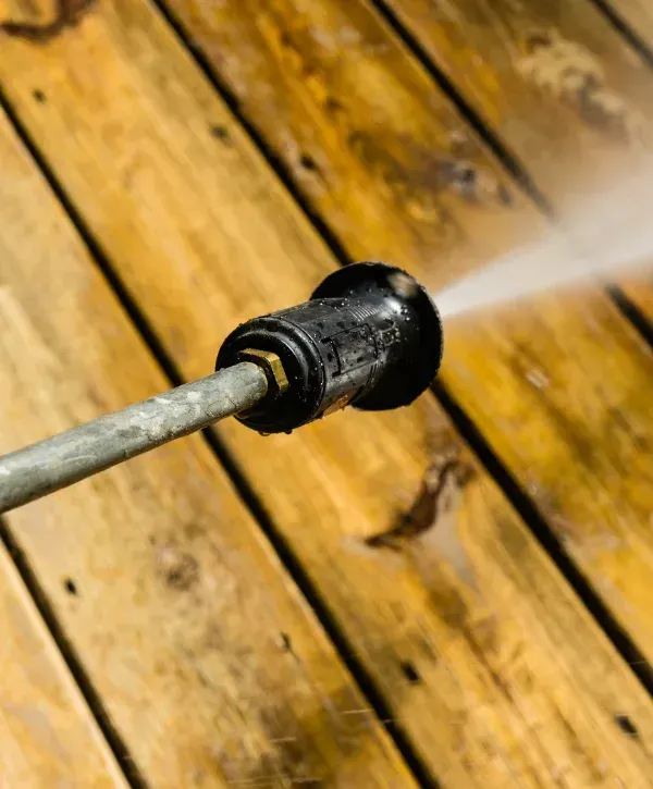 A close-up view of a power washer nozzle spraying water onto dirty wooden deck boards, cleaning them.