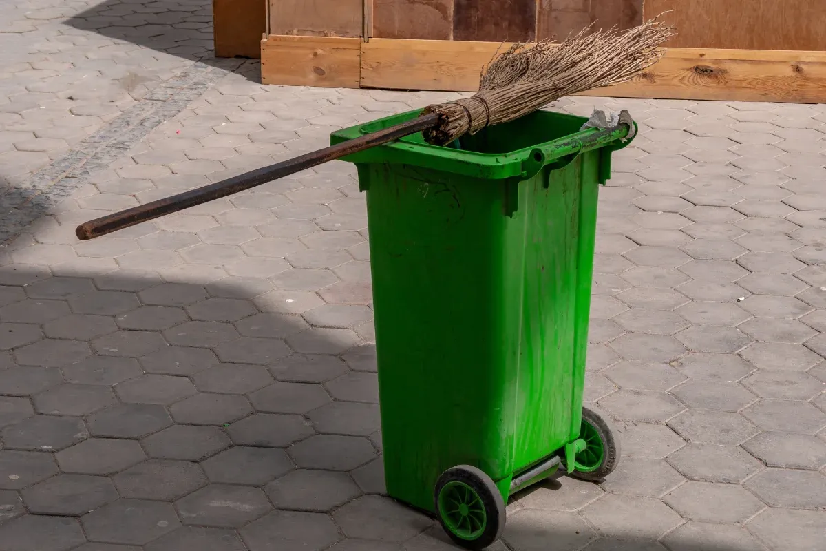 A bright green wheeled trash can with a traditional straw broom resting on its rim, on hexagonal paving stones.