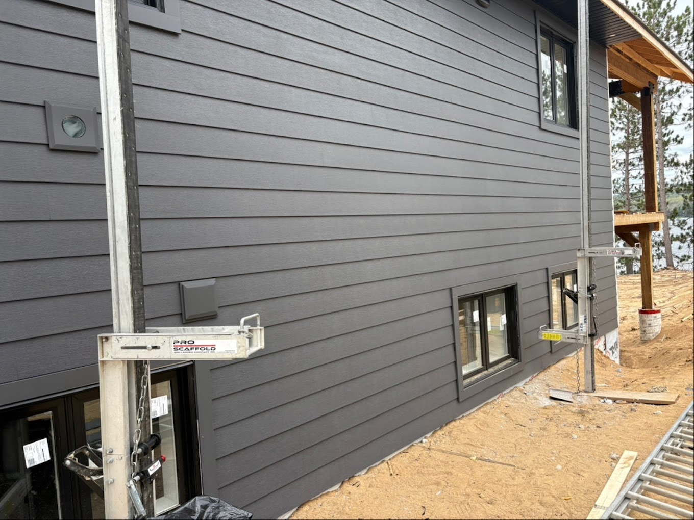 Exterior wall of a house featuring newly installed charcoal grey horizontal lap siding, framed windows, and matching utility vents, with metal Pro Scaffold pump jacks positioned on the dirt ground for installation work.