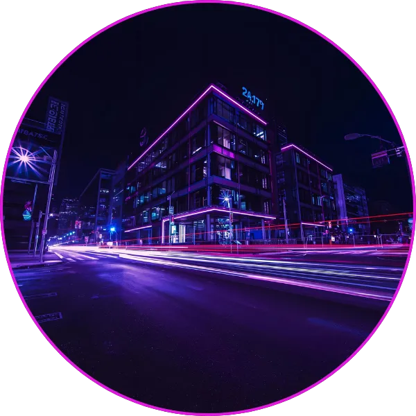 A nighttime cityscape with buildings outlined in purple neon lighting and long-exposure light trails from passing vehicles creating dynamic streaks across the street.