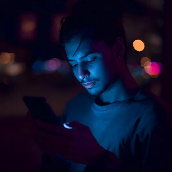 Man looking at a smartphone at night, his face illuminated by a blue glow with blurred city lights in the background.