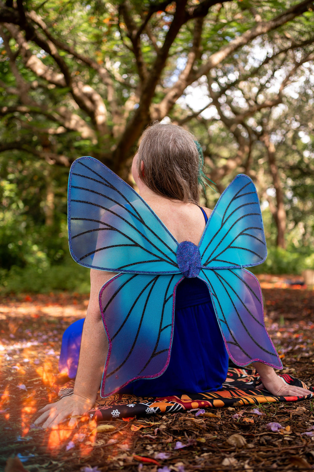 Woman in a long red dress wearing large translucent fairy wings, walking through a sunlit forest.