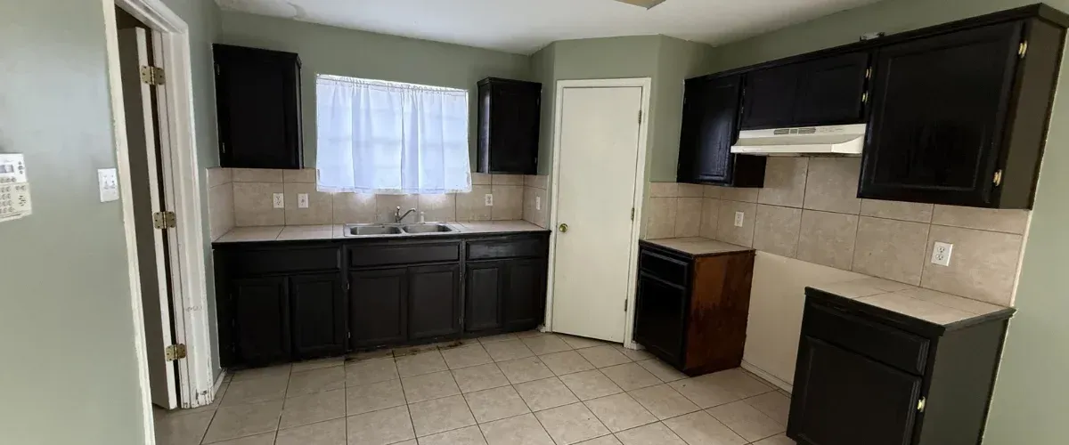 A dated kitchen with pale green walls, black painted cabinets, beige tile backsplashes and flooring, and a white sheer curtain.