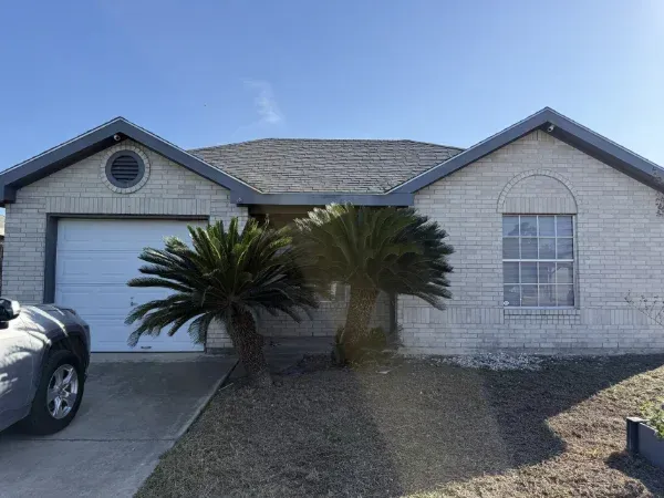A light brick-colored suburban house with a white garage door, two palm trees, and a parked car in the driveway.