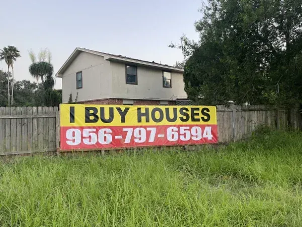 A yellow and red "I BUY HOUSES" banner with a phone number attached to a wooden fence, with tall grass in the foreground.