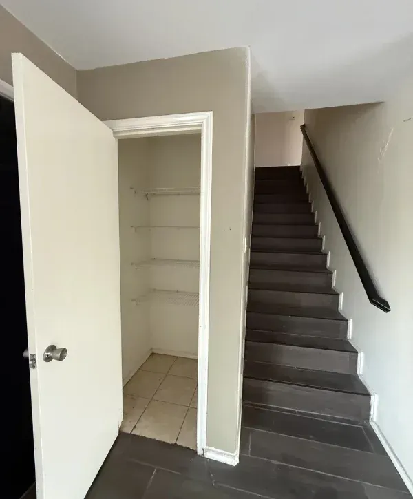 An interior view of an open closet under a dark staircase, featuring wire shelving and tiled floor