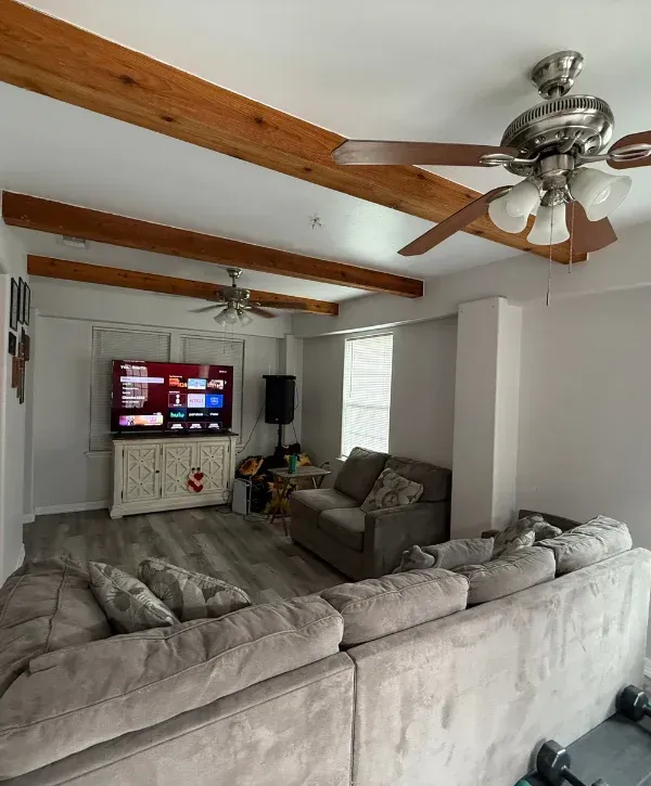 A cozy living room featuring light gray walls, wooden ceiling beams, a large TV, and comfortable sectional sofas.