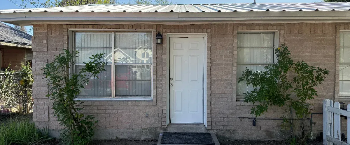 A close-up view of a light brown brick house with a white front door and two windows with blinds.