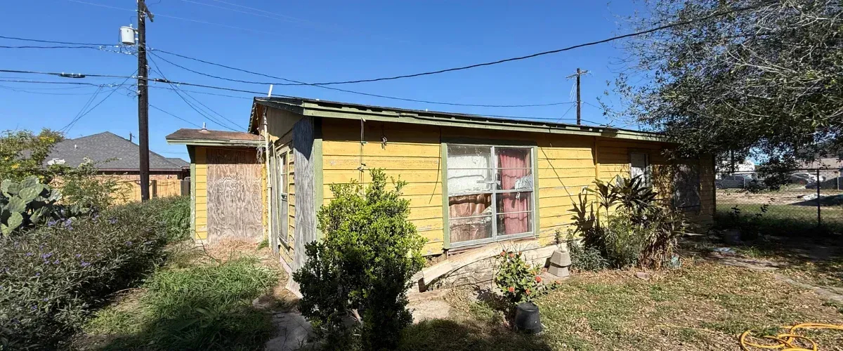 A dilapidated yellow house with peeling paint and boarded-up windows, surrounded by overgrown weeds under a clear blue sky.