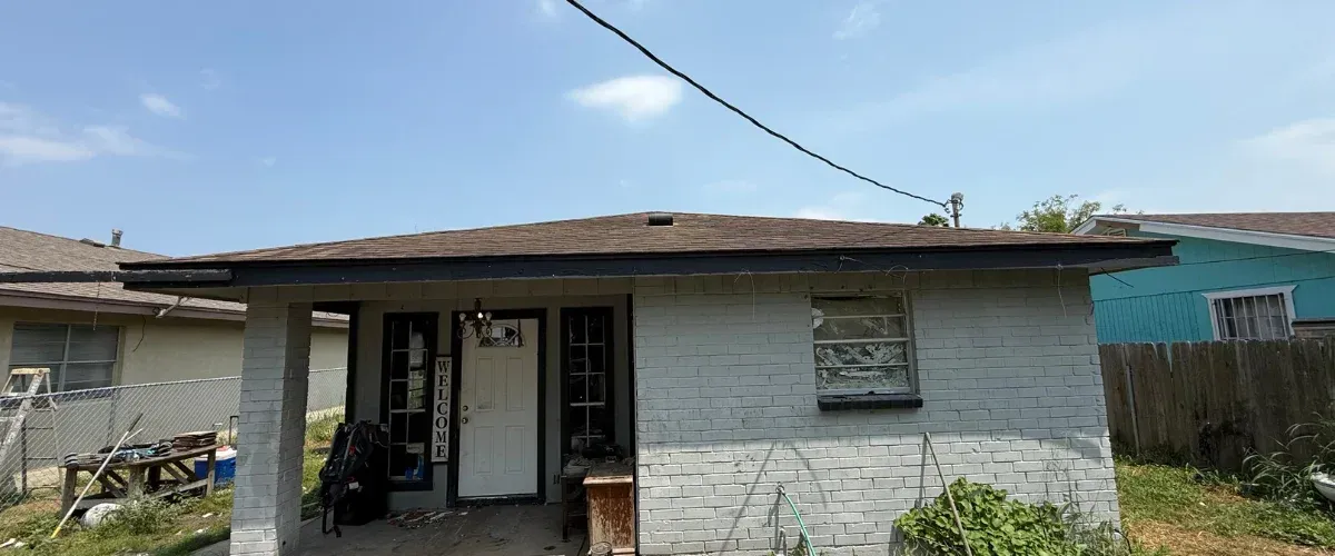 A partially dilapidated house with white brick, a dark roof, and a visible welcome sign by the front door on a sunny day.