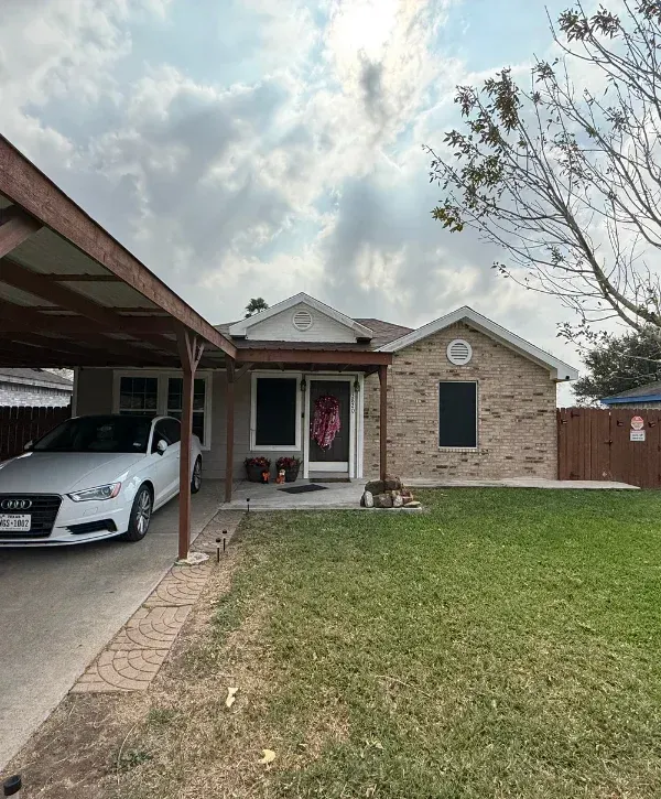 A white Audi sedan parked under a carport next to a brick house with a brown wooden fence on a cloudy day.