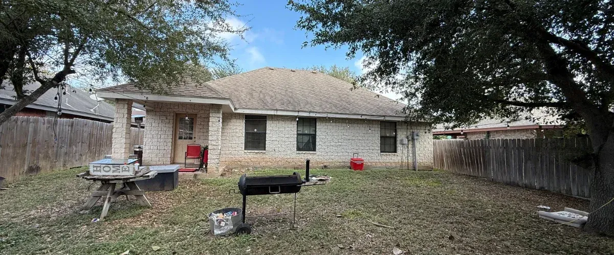 The back of a brick house with barred windows and an AC unit, set against a dry, brown grassy backyard on a sunny day.