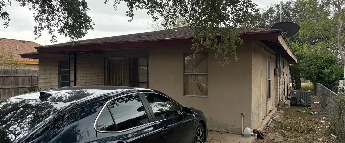 A single-story house with a worn roof and stucco walls, partially obscured by trees, with a black car parked in front.