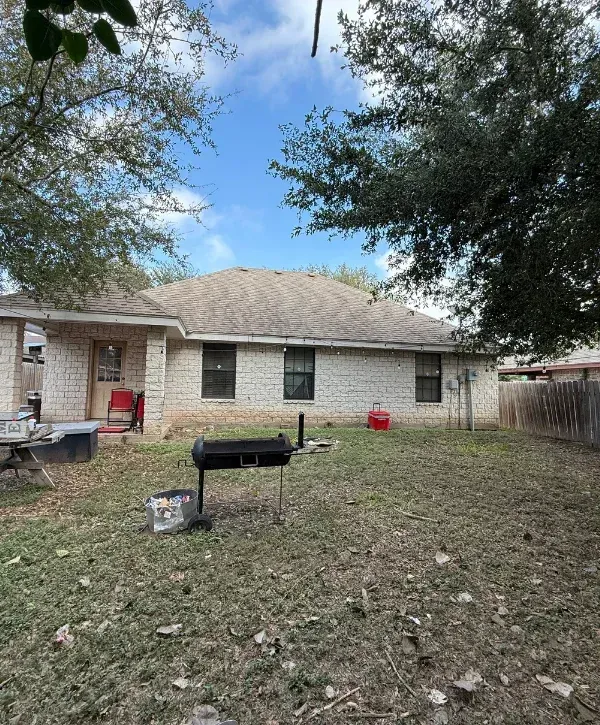 Rear view of a light brick house with a brown roof, grill, trash can, and red cooler in the backyard under a blue sky.